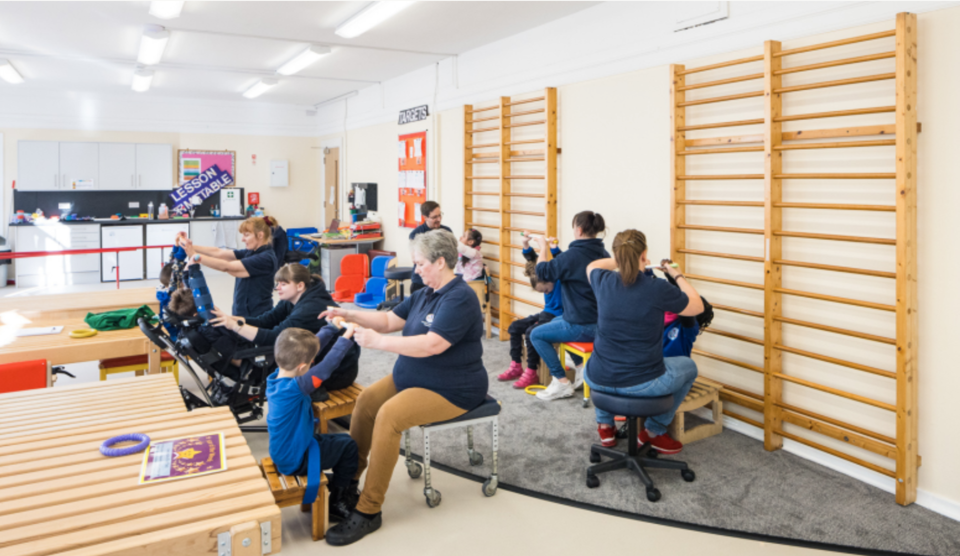 A group of adults and children in a brightly lit classroom setting. The adults are assisting the children with various activities, some of whom are using adaptive equipment. Wooden climbing bars are mounted on the wall, and there are benches, tables, and colourful educational tools in the background.