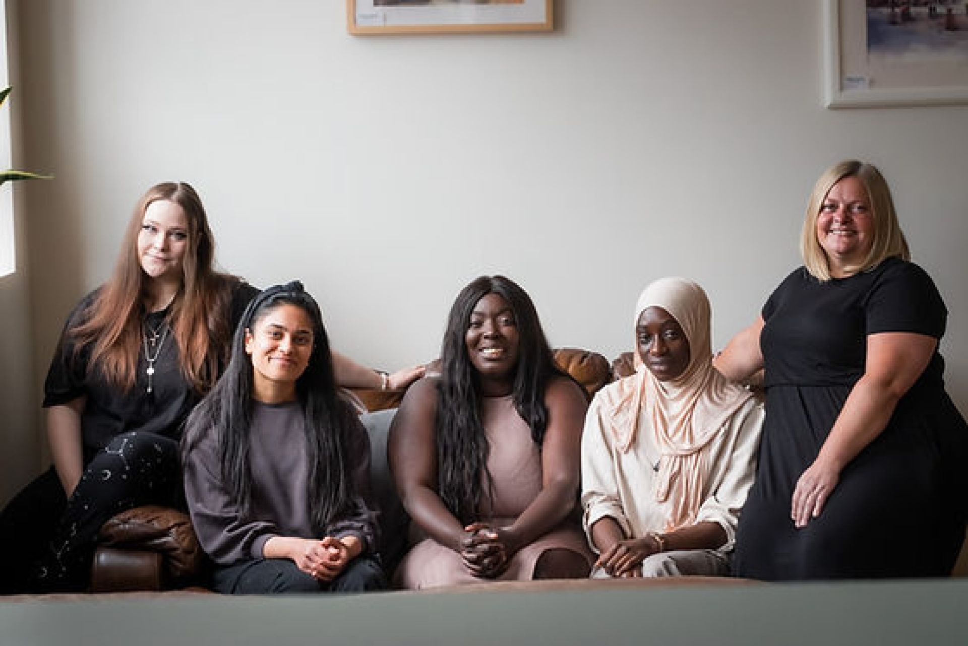 A group of five women sitting together on a brown leather couch in a warmly lit room.