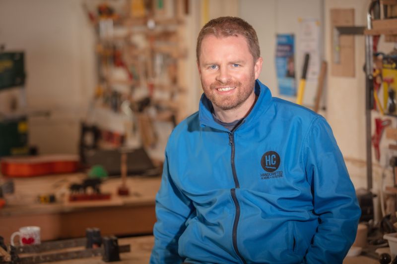 The image shows a man smiling in a workshop environment. He is wearing a blue jacket with the logo "HC Handcrafted" on the left side of his chest. The workshop is filled with various tools and equipment in the background. The environment appears to be focused on creating or repairing items.