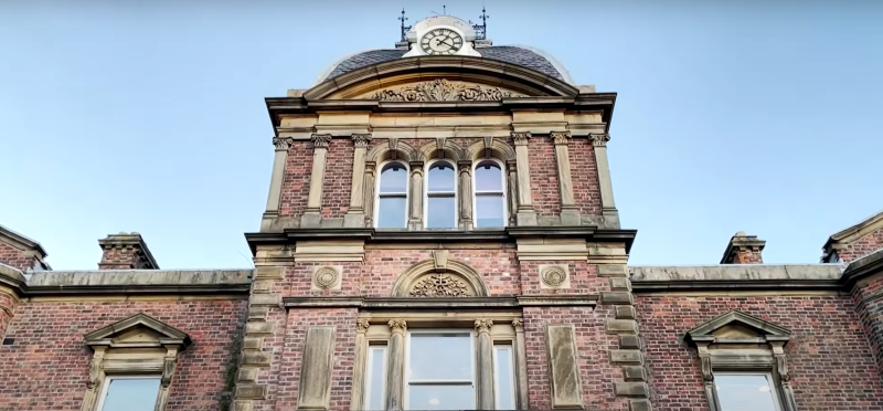 An ornate red brick building with Victorian-style architecture, featuring a central clock tower, arched windows, and intricate stone detailing. The structure exudes historical charm, framed against a clear blue sky.