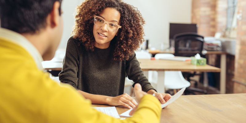Woman looks at a sheet of paper while sat at a desk with the back of a person facing her
