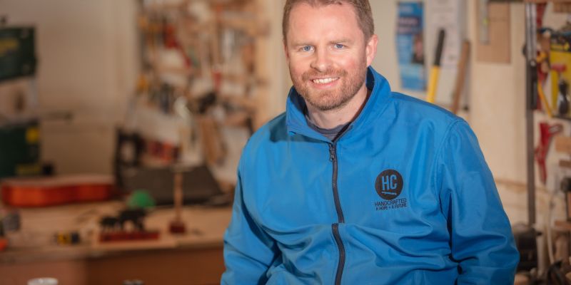 The image shows a man smiling in a workshop environment. He is wearing a blue jacket with the logo "HC Handcrafted" on the left side of his chest. The workshop is filled with various tools and equipment in the background. The environment appears to be focused on creating or repairing items.