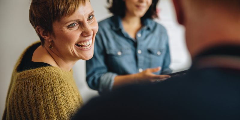 The image shows a woman with short, light brown hair wearing a mustard-coloured jumper, smiling as she engages in conversation. In the background, another woman with dark hair, dressed in a denim shirt, looks on with a gentle expression. The scene appears casual and friendly, with a relaxed atmosphere.