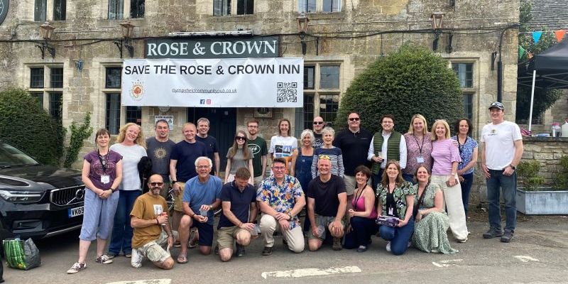 Picture showing a number of people looking ahead. There is an old rustic building behind them with a banner that says 'ROSE & CROWN'