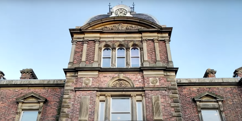 An ornate red brick building with Victorian-style architecture, featuring a central clock tower, arched windows, and intricate stone detailing. The structure exudes historical charm, framed against a clear blue sky.