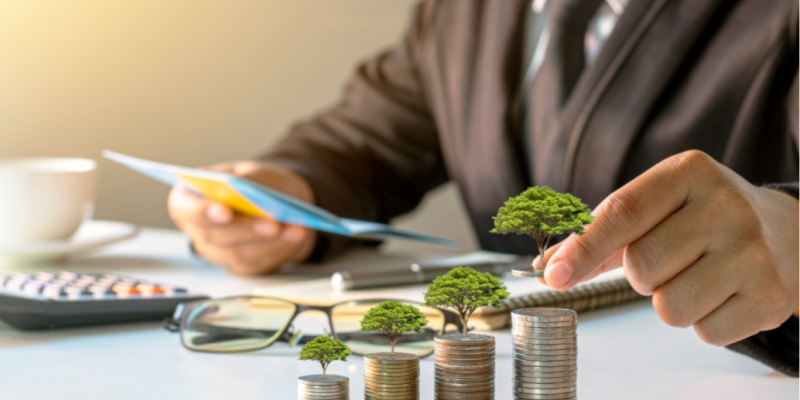 A business professional in a suit is seated at a desk, holding documents. In the foreground, stacks of coins gradually increase in height, with each stack featuring a small tree growing on top. A calculator, glasses, and a cup of coffee are also visible on the desk.