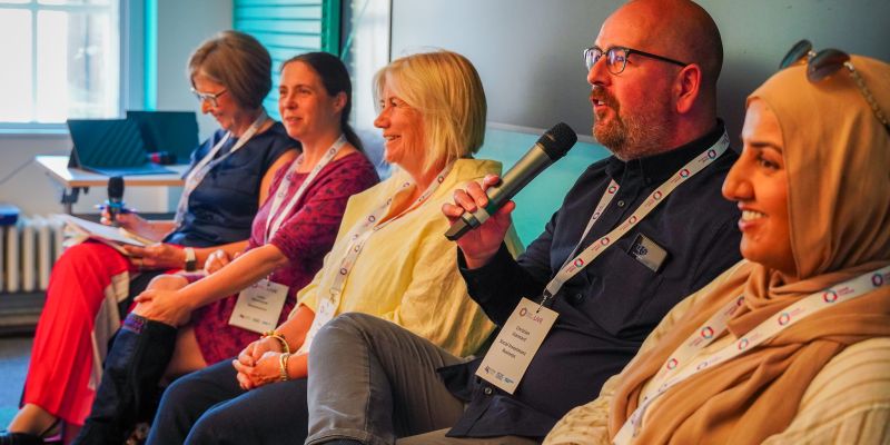 A panel of 5 people sitting, a man is holding the microphone